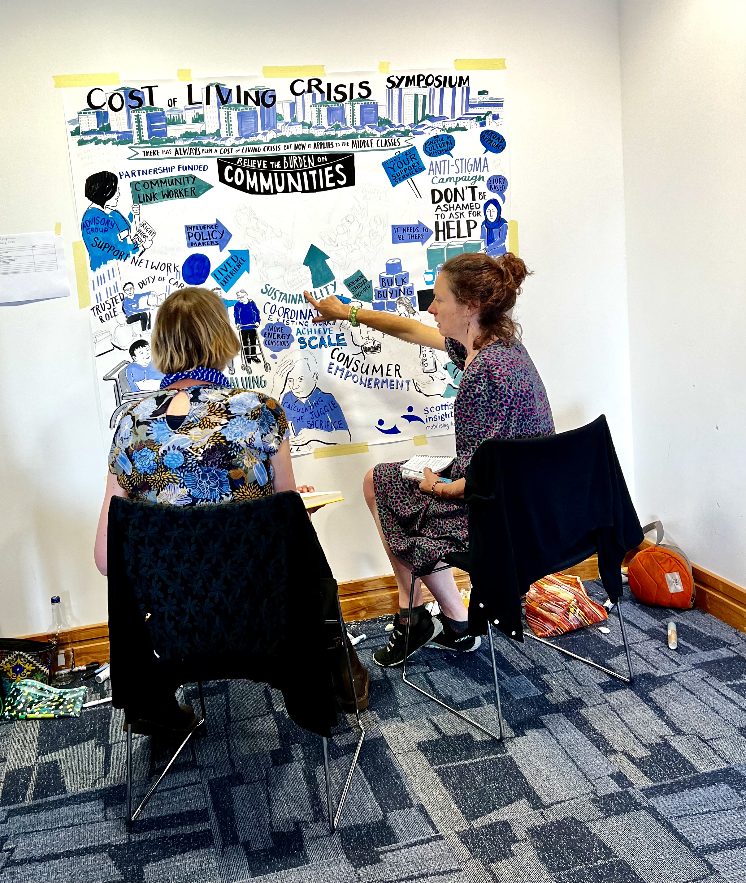Two artists are sitting facing a large sheet of paper on the wall, showing a work in progress of their illustration.